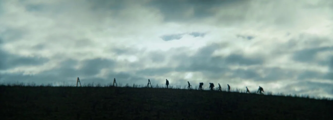 28 Years Later (2025) movie still: wide — A dark, grassy hill rises against a heavy, overcast sky filled with clouds. A group of people walks in a…