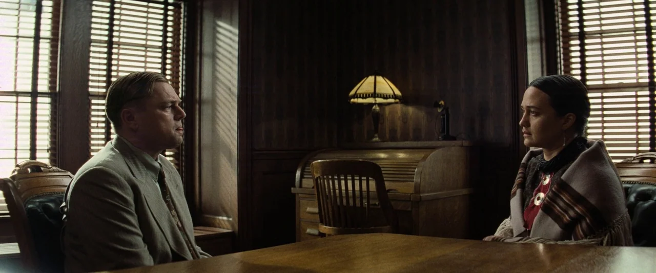 Killers of the Flower Moon (2023) movie still: medium — A dark, wood-paneled office space features two windows with closed wooden blinds. A man and…