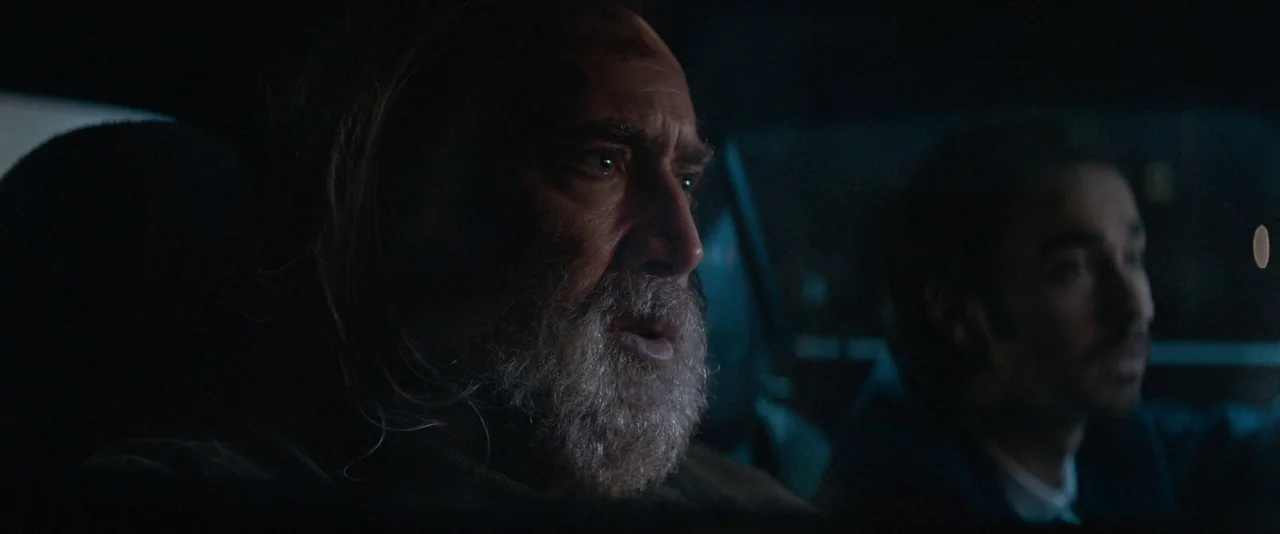 Pig (2021) movie still: closeup — An older man and a younger man sit inside the cabin of a dark vehicle. The interior is dimly lit by cool light…