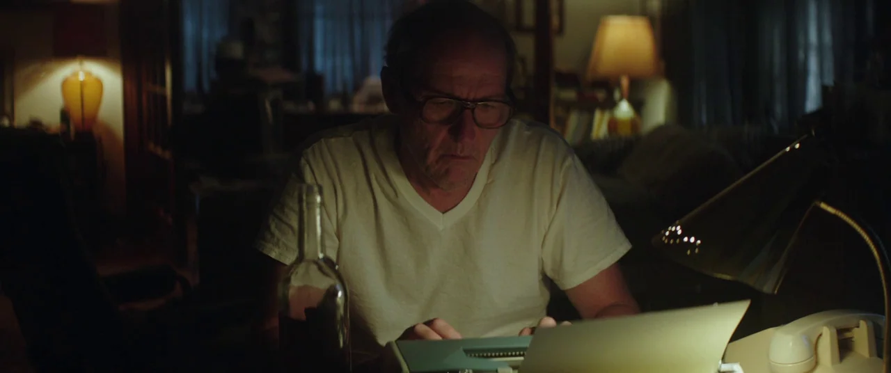 God’s Pocket (2014) movie still: medium — A dimly lit home study contains a typewriter, a bottle, and a telephone. A man wearing glasses sits at the…