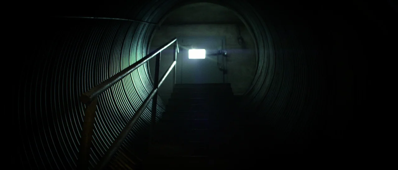 10 Cloverfield Lane (2016) movie still: establishing — A dark, cylindrical corrugated metal tunnel leads upward to a closed steel door. A stark…