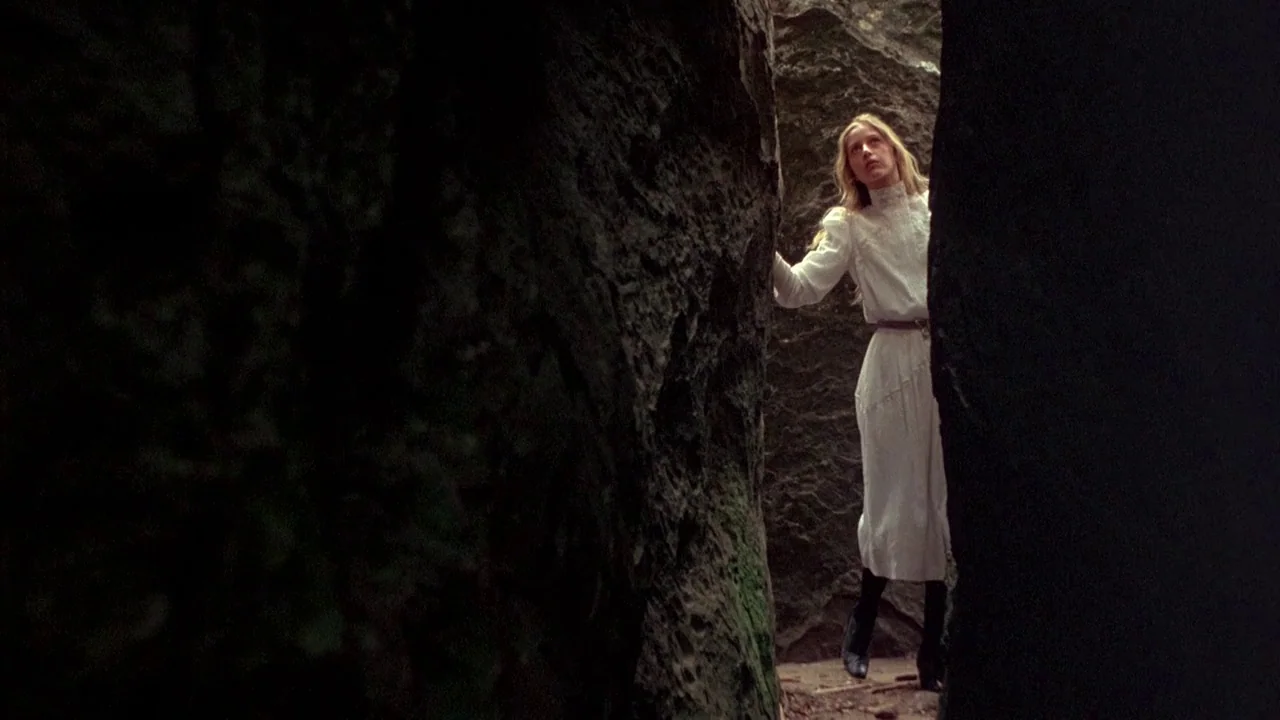 Picnic at Hanging Rock (1975) movie still: fullbody — A woman stands between two massive, dark rock faces in a rugged outdoor setting. The woman in…