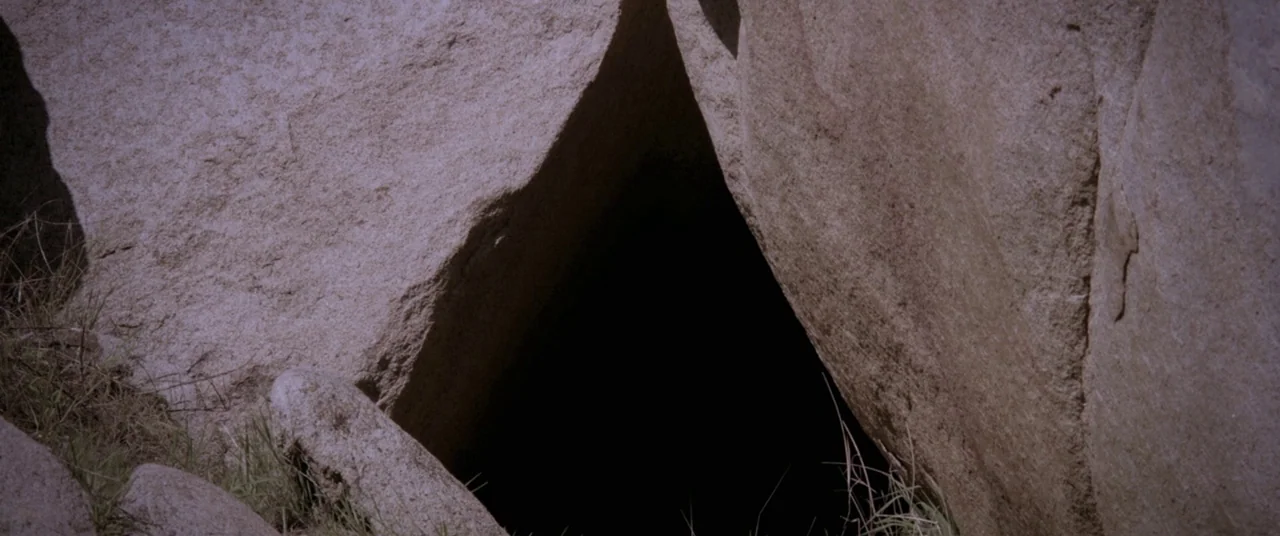Here Comes The Devil (2012) movie still: closeup — A dark triangular opening is formed by large, light-colored jagged rock formations. Sparse green…