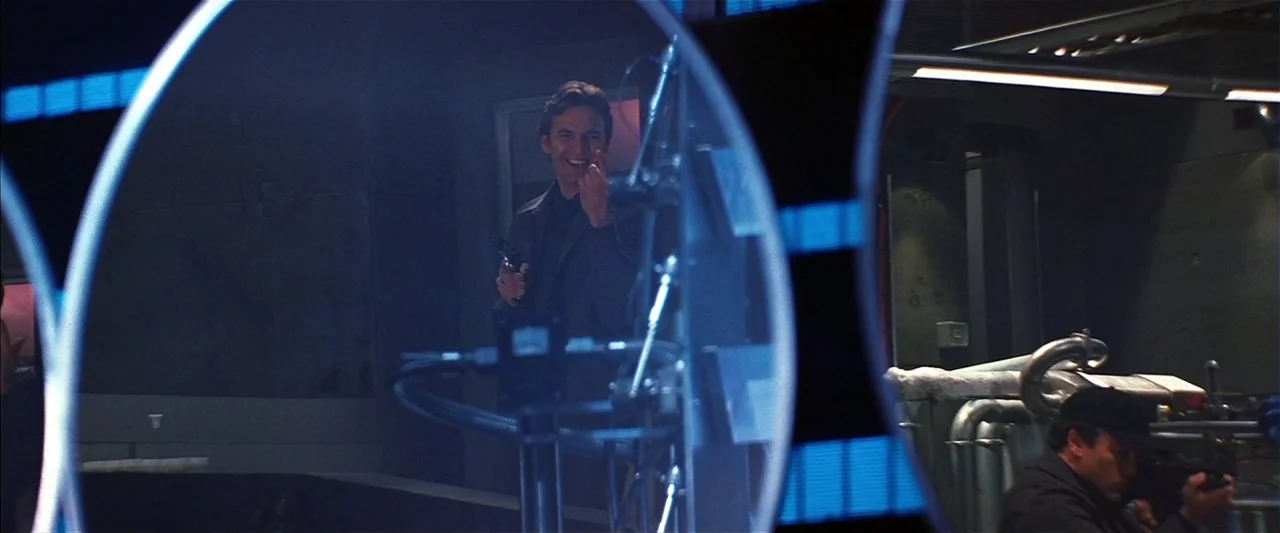 Mission: Impossible II (2000) movie still: medium — A dimly lit industrial room features complex metal piping and machinery in the foreground. A man…