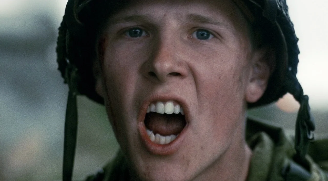 Saving Private Ryan (1998) movie still: closeup — A soldier in uniform stands on a battlefield with an overcast sky behind him. He shouts with his…