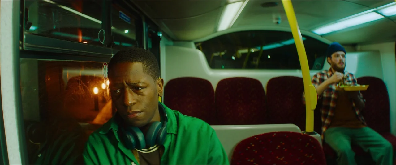 Rye Lane (2023) movie still: medium — The interior of a public bus at night has red patterned seats and yellow poles. A man with headphones sits in…