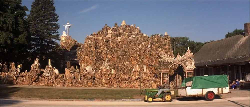The Straight Story (1999) movie still: wide — An outdoor religious grotto features intricate stone and rock formations with a crucifix statue. A man…