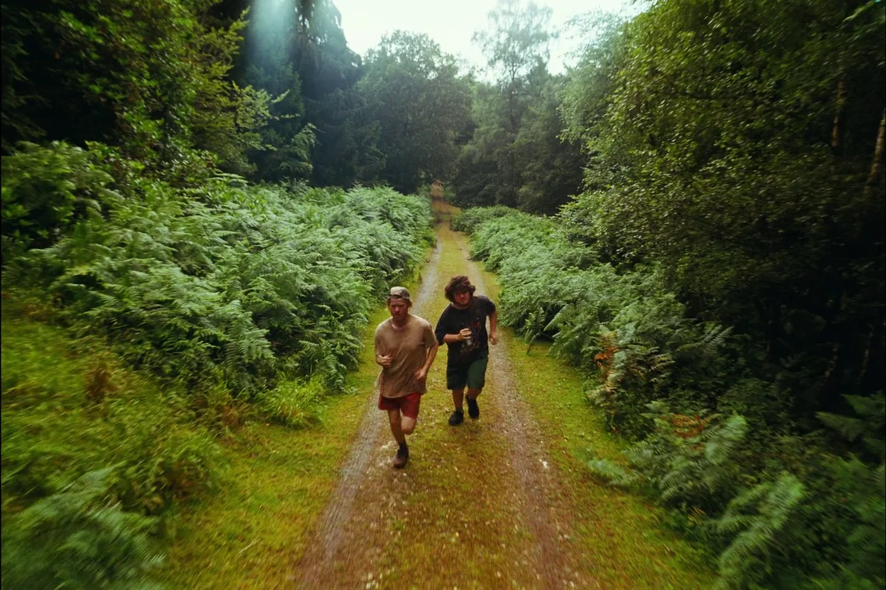Bugonia (2025) movie still: fullbody — A narrow dirt path winds through a dense woodland area filled with tall ferns. Two young men run along the…