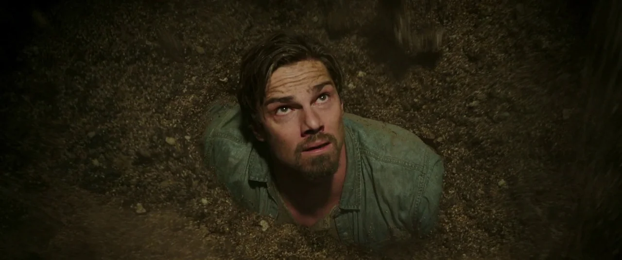 It Chapter Two (2019) movie still: closeup — A man is buried up to his chest in a dark, dirt-filled pit. He looks upward with a distressed…