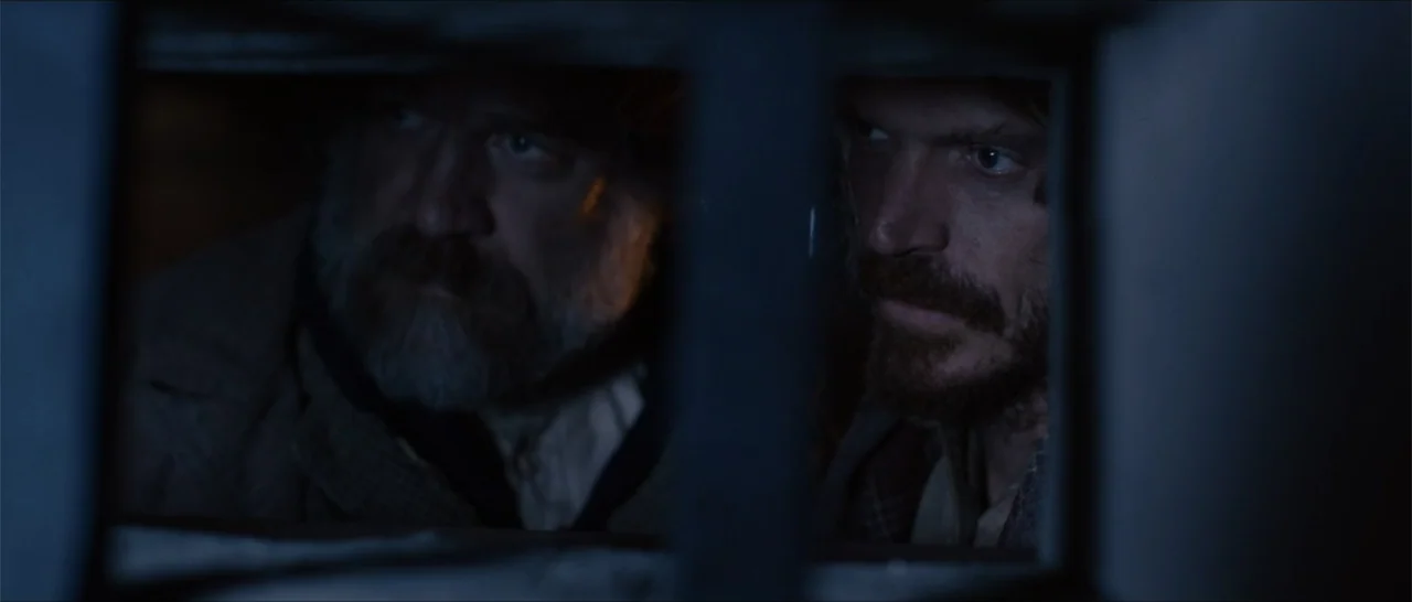 Out of Liberty (2019) movie still: medium — Two men look through vertical metal bars in a dark, confined space. The men peer intensely at something…