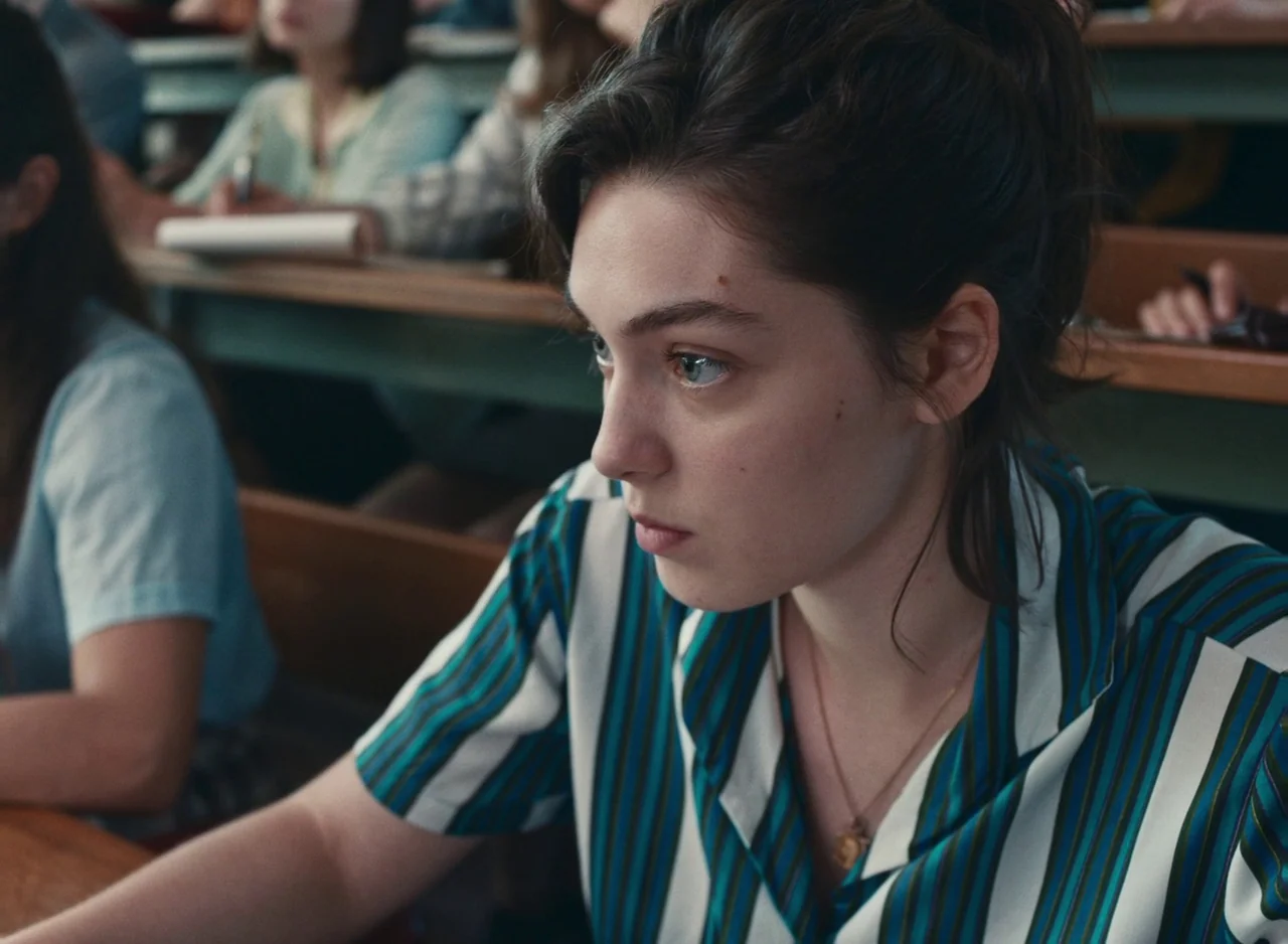 Happening (2021) movie still: medium — A crowded university lecture hall features rows of wooden desks with green-painted supports. A young woman…
