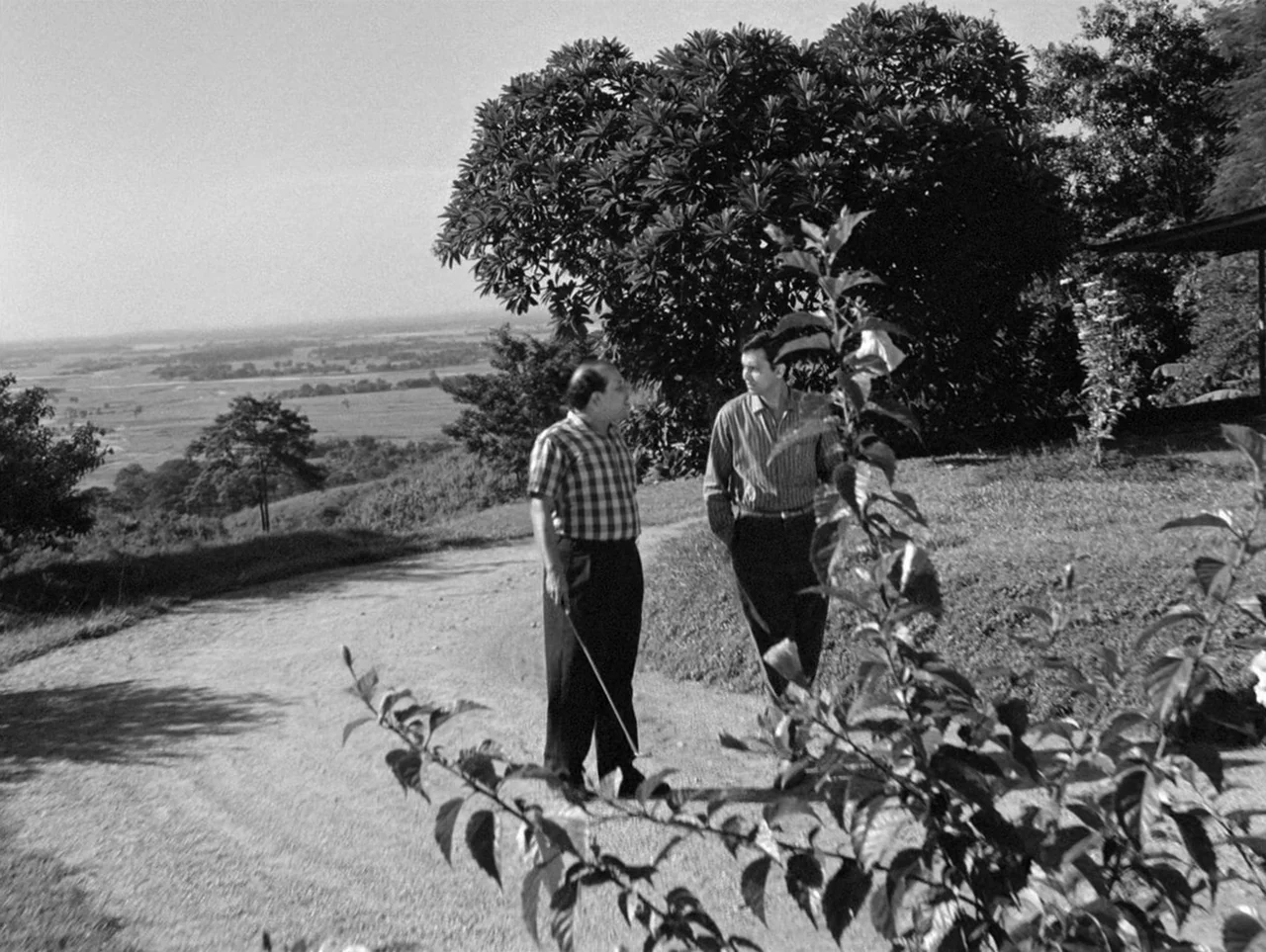 The Coward (1965) movie still: fullbody — A dirt path cuts through a grassy hill overlooking a vast rural valley landscape. Two men in collared…