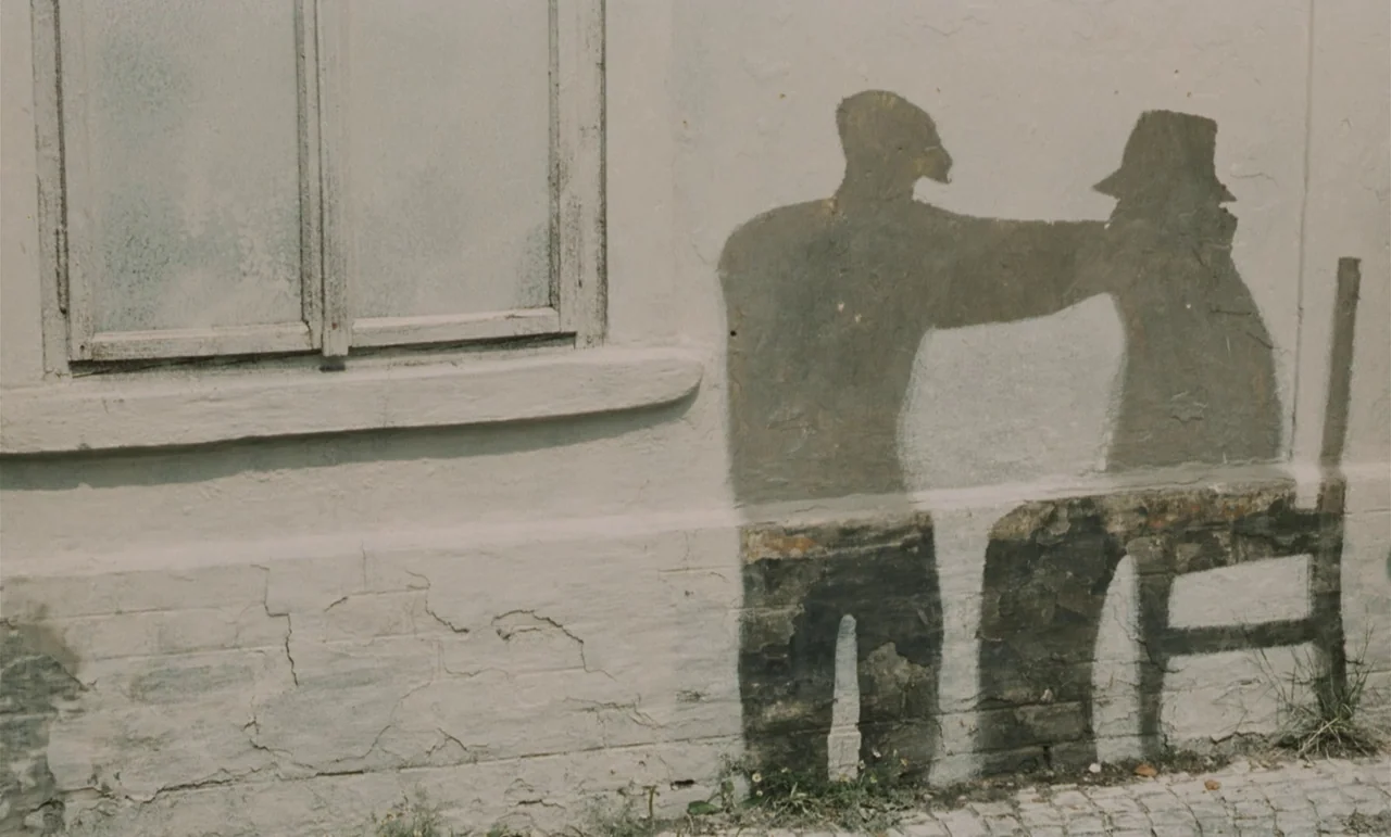 Larks On A String (1969) movie still: medium — A light-colored brick wall features a weathered, painted mural of two figures standing together. A…