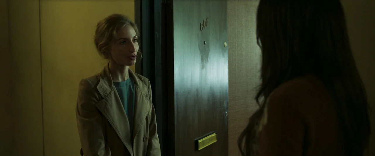 Stopmotion (2024) movie still: medium — An apartment hallway with a brown door numbered 801 in metallic digits. A woman in a trench coat faces an…