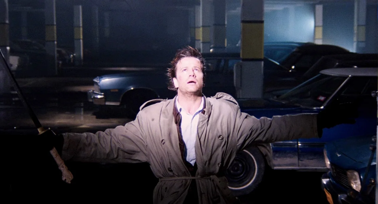 Highlander (1986) movie still: medium — A dimly lit concrete parking structure with parked cars and support pillars. A man wearing a tan trench coat…