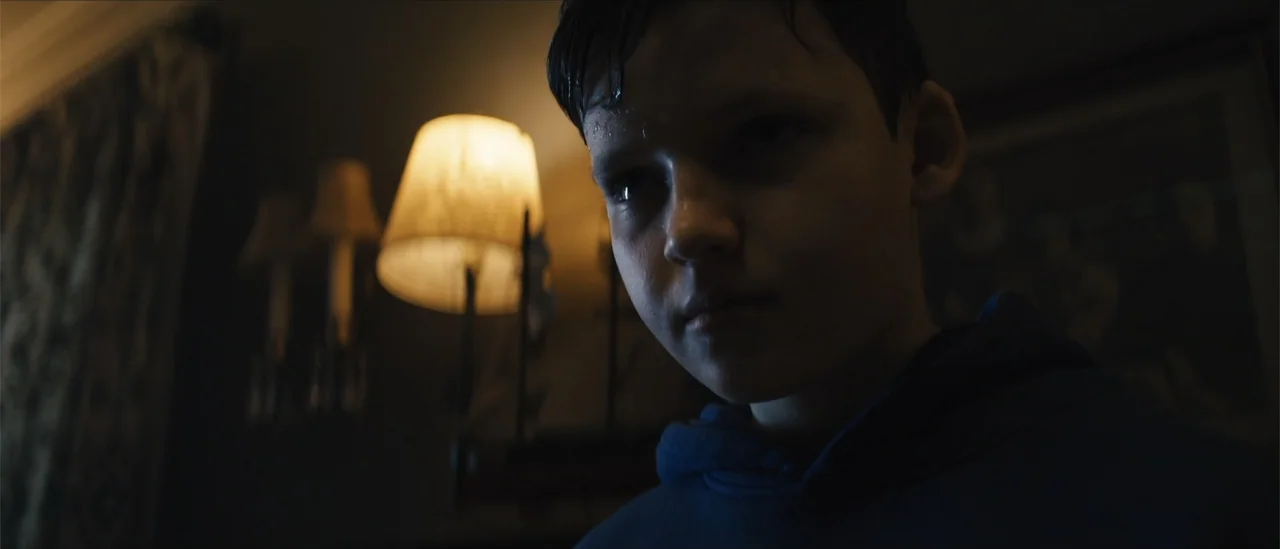 All Fun and Games (2023) movie still: closeup — A dimly lit room features a glowing lampshade in the background behind a subject. A young boy with…