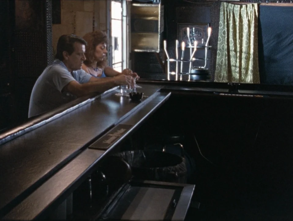 River of Grass (1994) movie still: medium — A dimly lit bar features a long dark wooden counter and patterned window curtains. A man and a woman sit…