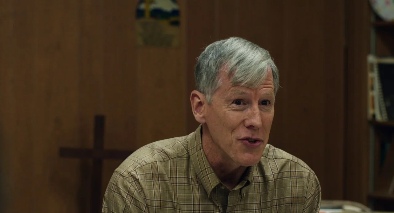 The Miseducation of Cameron Post (2018) movie still: closeup — A man stands in an office with dark wood paneling and a wall-mounted cross. The man…
