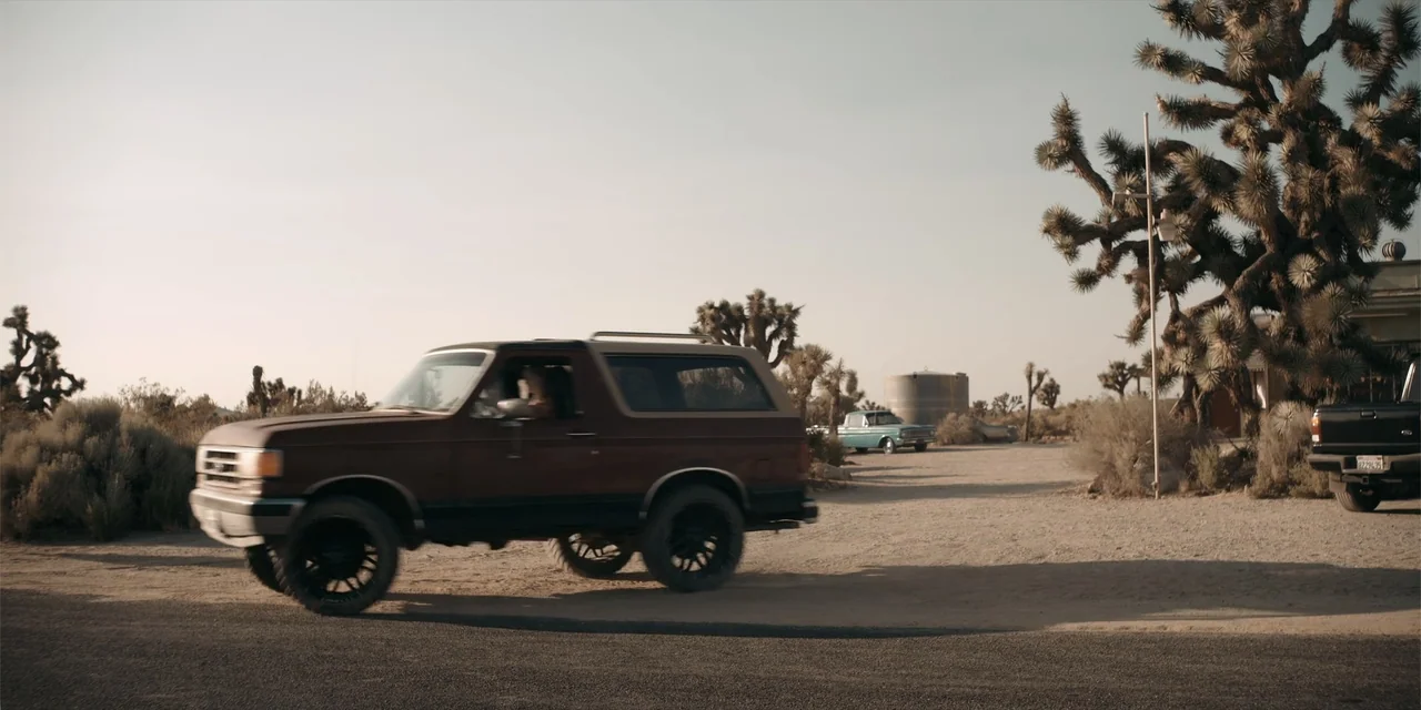 Fall (2022) movie still: wide — A dry desert landscape features Joshua trees and dirt terrain under a clear sky. A dark-colored SUV moves across the…