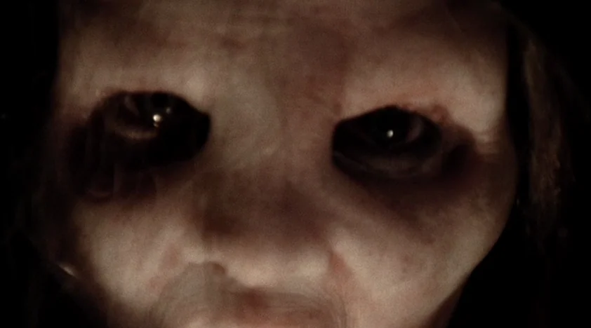 The House Of The Devil (2009) movie still: closeup — The face of a pale figure emerges from absolute darkness. The subject stares forward with dark,…