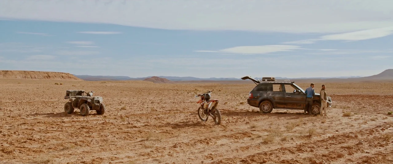 Revenge (2017) movie still: wide — A vast, dry desert landscape stretches out under a wide, pale blue sky. Two men stand beside their mud-caked…