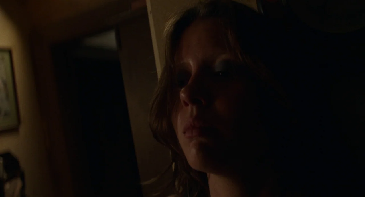 X (2022) movie still: closeup — A dimly lit narrow hallway with a dark wall and a framed picture. A young woman stands in the shadows looking…