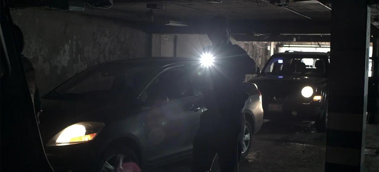 About the Deaths (2016) movie still: medium — A dimly lit concrete parking structure with two parked vehicles flanking the central figure. A person…