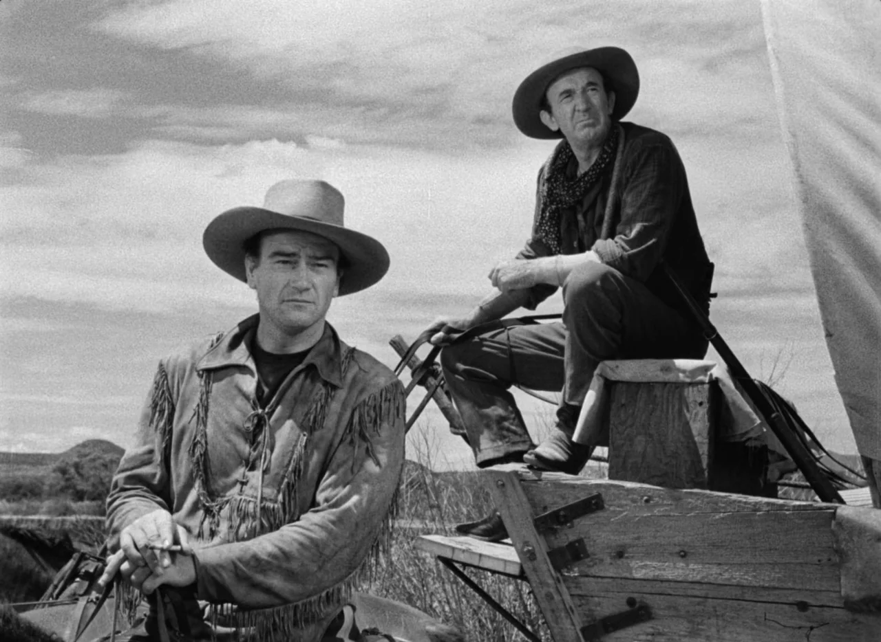 Red River (1948) movie still: medium — A vast, open prairie under a clouded sky features a wooden pioneer wagon. Two men in western gear sit near…