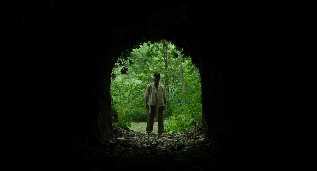 The Mimic (2017) movie still: wide — A dark, arched cave mouth opens onto a lush green forest filled with foliage. A man in light-colored clothing…