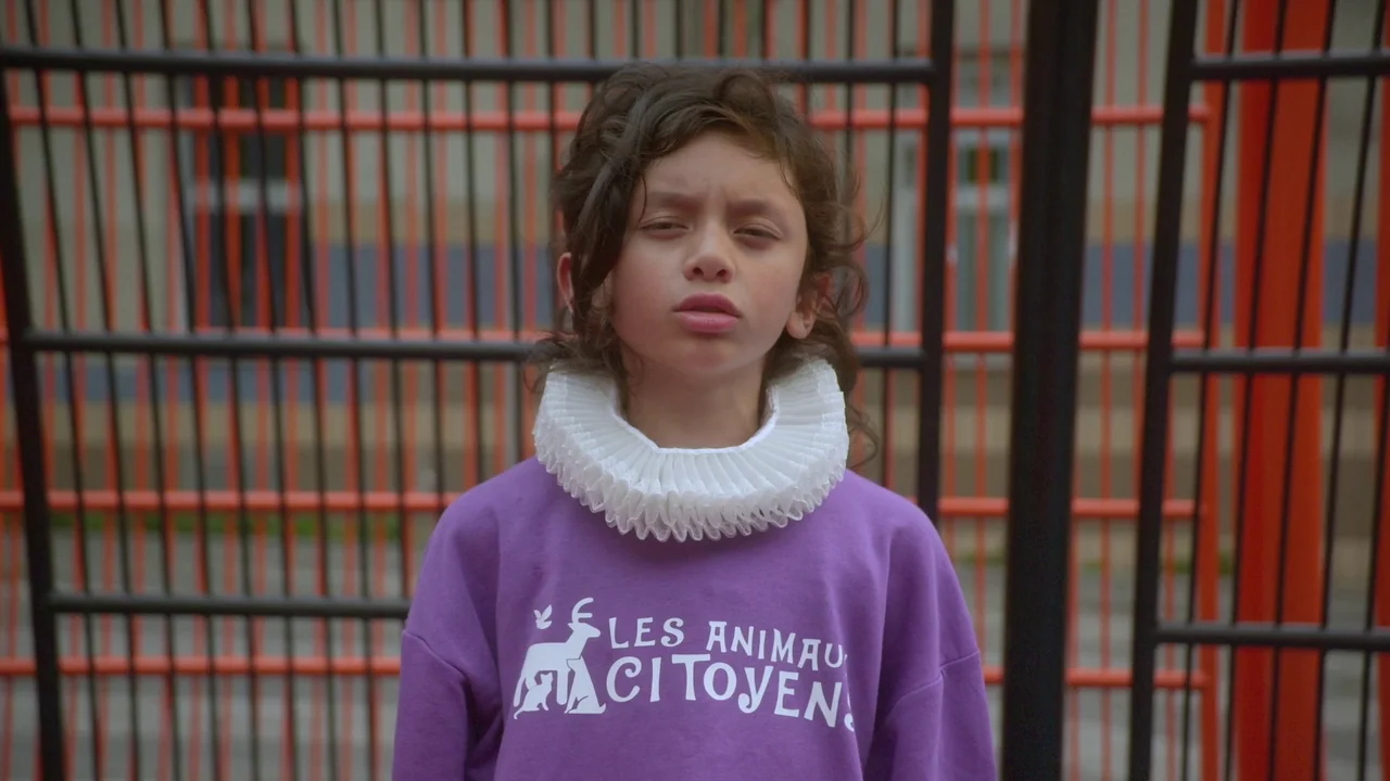 Orlando: My Political Biography (2023) movie still: medium — An outdoor area with a vertical black and orange grid fence background. A child wearing…