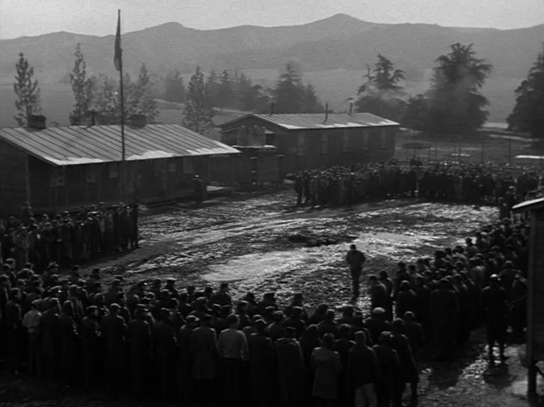 Stalag 17 (1953) movie still: establishing — A military camp courtyard with wooden barracks and a flagpole surrounded by muddy ground. A large group…
