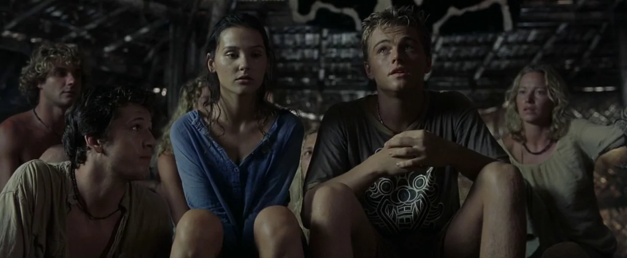 The Beach (2000) movie still: medium — A dark wooden hut interior features a group of young adults sitting together closely. They gaze toward an…