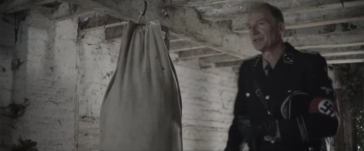 Behind the Line: Escape to Dunkirk (2020) movie still: medium — A basement space has exposed wooden ceiling beams and rough, whitewashed brick…
