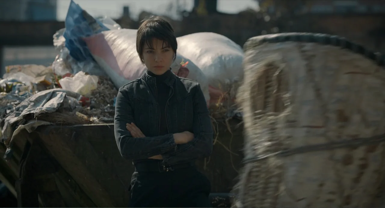 Ice Road: Vengeance (2025) movie still: medium — An outdoor area cluttered with large bags of refuse in a metallic industrial container. A young…