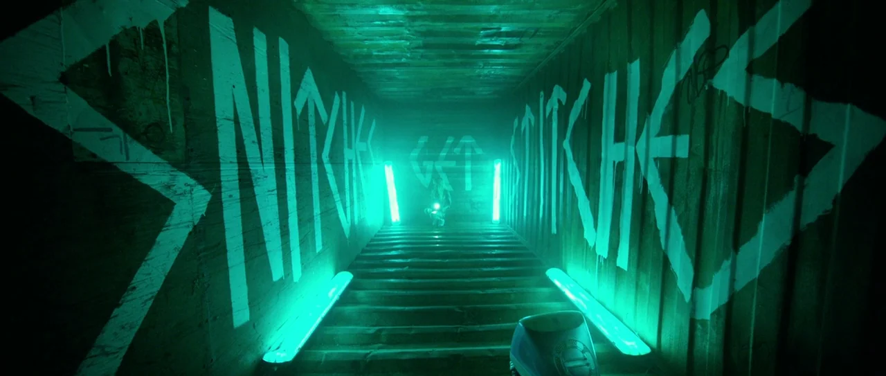 Nerve (2016) movie still: wide — A dark, narrow metal shipping container features white graffiti text on both corrugated walls. A person stands at…