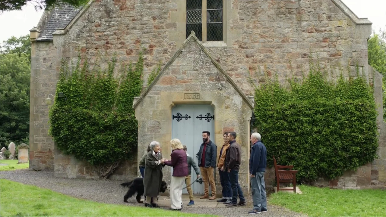 A Scottish Love Scheme (2024) movie still: wide — A group of six people stand before the blue wooden doors of a stone building. The structure…