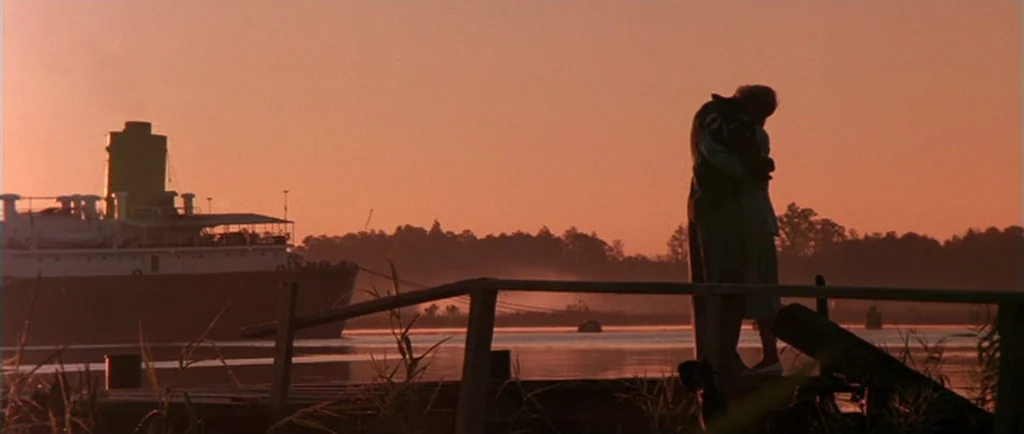 Manhunter (1986) movie still: wide — A wooden pier extends into calm water under a soft orange sunrise sky. Two people stand embracing on the pier…