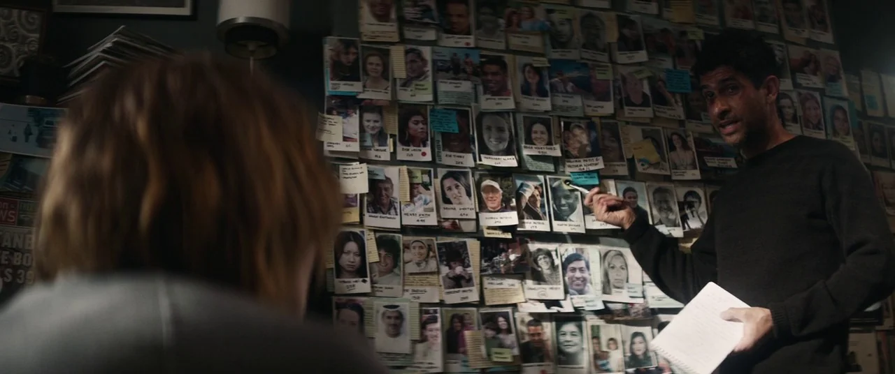 The Rhythm Section (2020) movie still: medium — An interior space features a wall densely covered with numerous printed portrait photographs. A man…