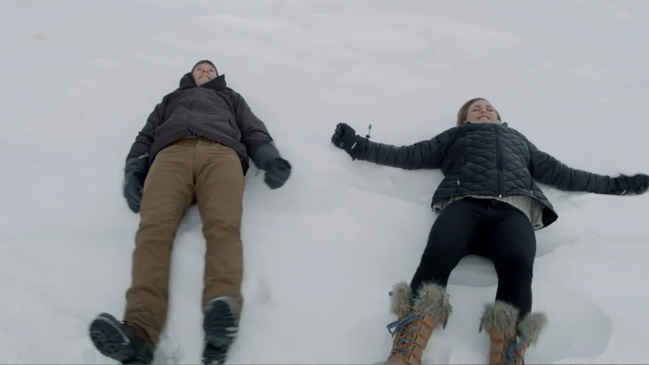 Alex & Emma (2003) movie still: fullbody — An expanse of untouched white snow covering the ground during an overcast day. A man and a woman lie flat…