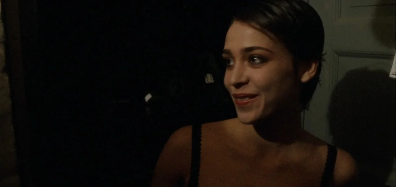 Pusher (1996) movie still: medium — A dark indoor setting with limited illumination on the subject's face. A woman with short hair turns her head…
