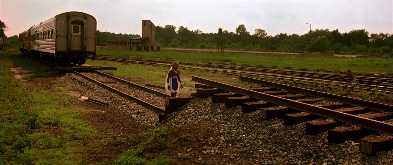 George Washington (2000) movie still: wide — A rural railway yard features rusted metal tracks and a silver train car. A small child in a makeshift…