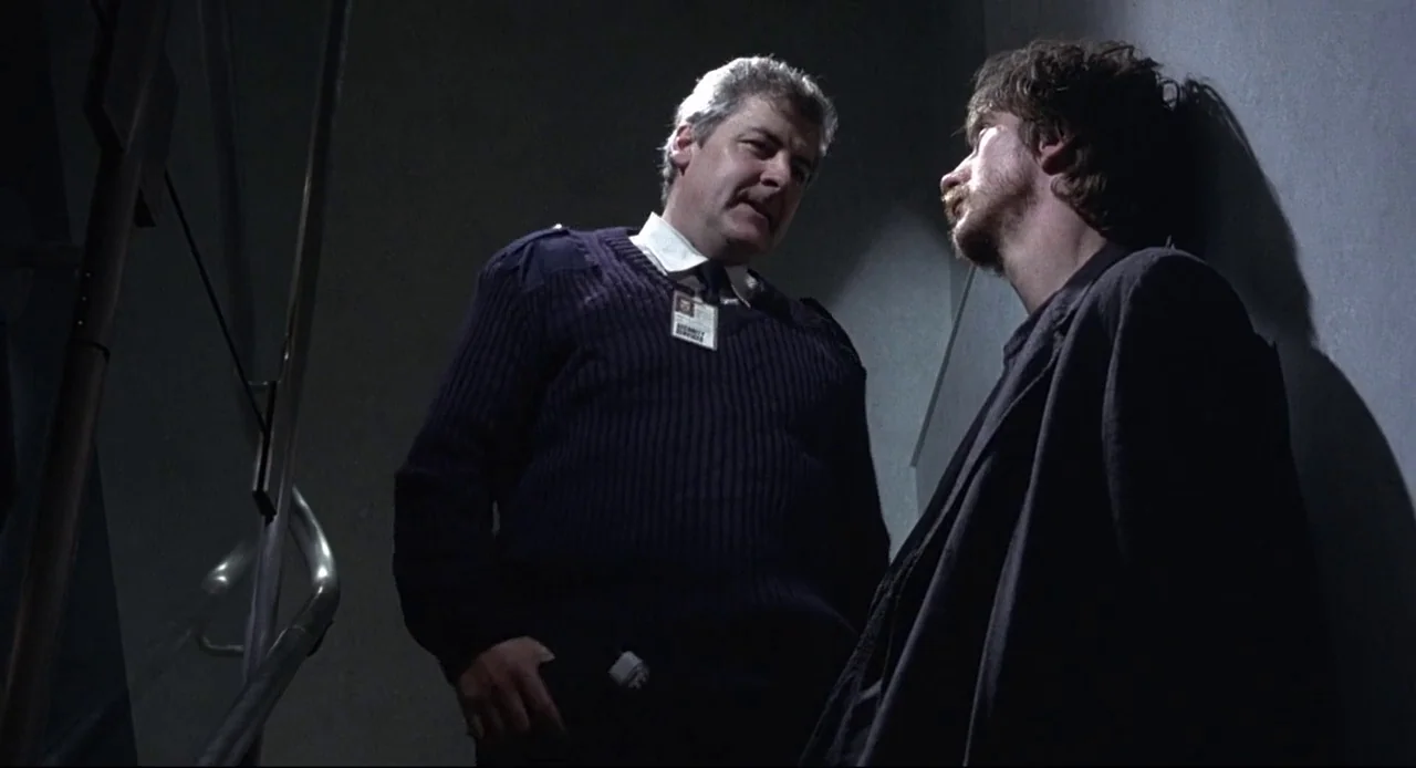 Naked (1993) movie still: medium — An industrial stairwell with concrete walls and a metal handrail in dim lighting. A uniformed security guard…