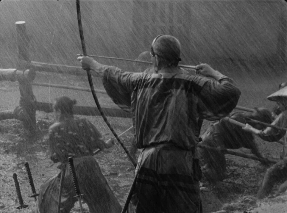 Seven Samurai (1954) movie still: medium — A wet, muddy outdoor area features a wooden rail fence in heavy rain. A samurai turns his back to the…