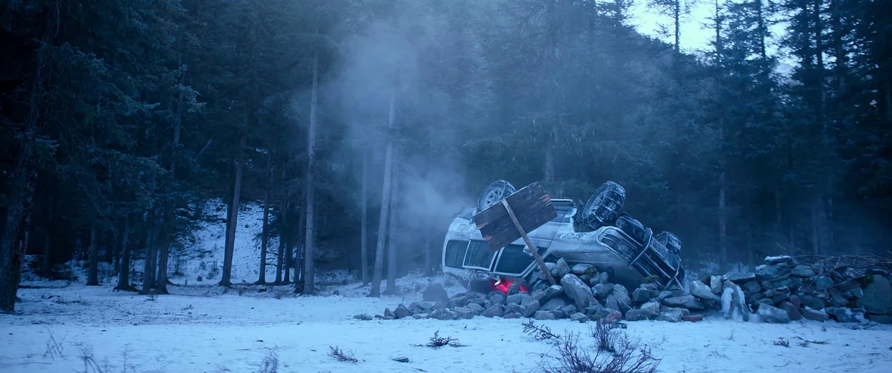 One And Four (2021) movie still: wide — A white SUV lies overturned against a pile of large rocks in a snow-covered forest. Smoke billows from the…