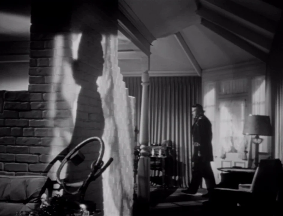 The Stranger (1946) movie still: wide — A dark, mid-century living room features a prominent painted brick pillar and heavy curtains. A man in a…
