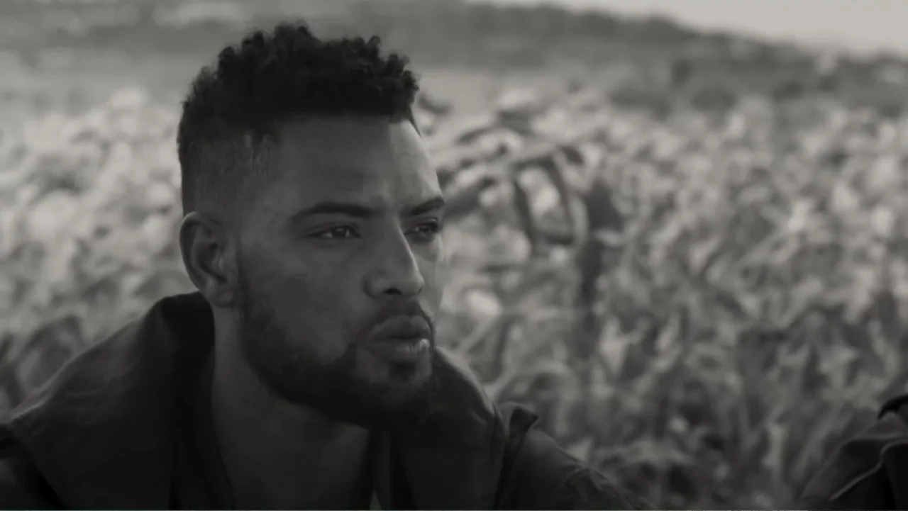 Faya Dayi (2021) movie still: closeup — A field of crops stretches into the out-of-focus background on a bright day. A young man with a short beard…