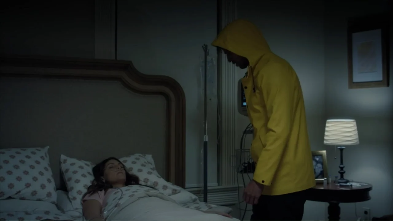 Storm Damage (2000) movie still: medium — A dimly lit room containing a bed and medical equipment near a small table. A man wearing a yellow hooded…