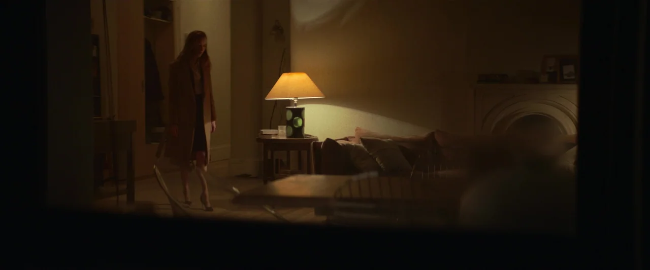 Fair Play (2023) movie still: wide — A dimly lit living room features a table lamp casting light on a couch. A woman wearing a long coat stands in…