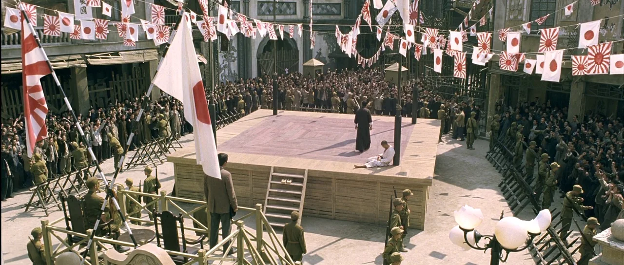 Ip Man (2008) movie still: wide — An open square filled with many people and rows of flags above a wooden stage. A man in black stands over another…
