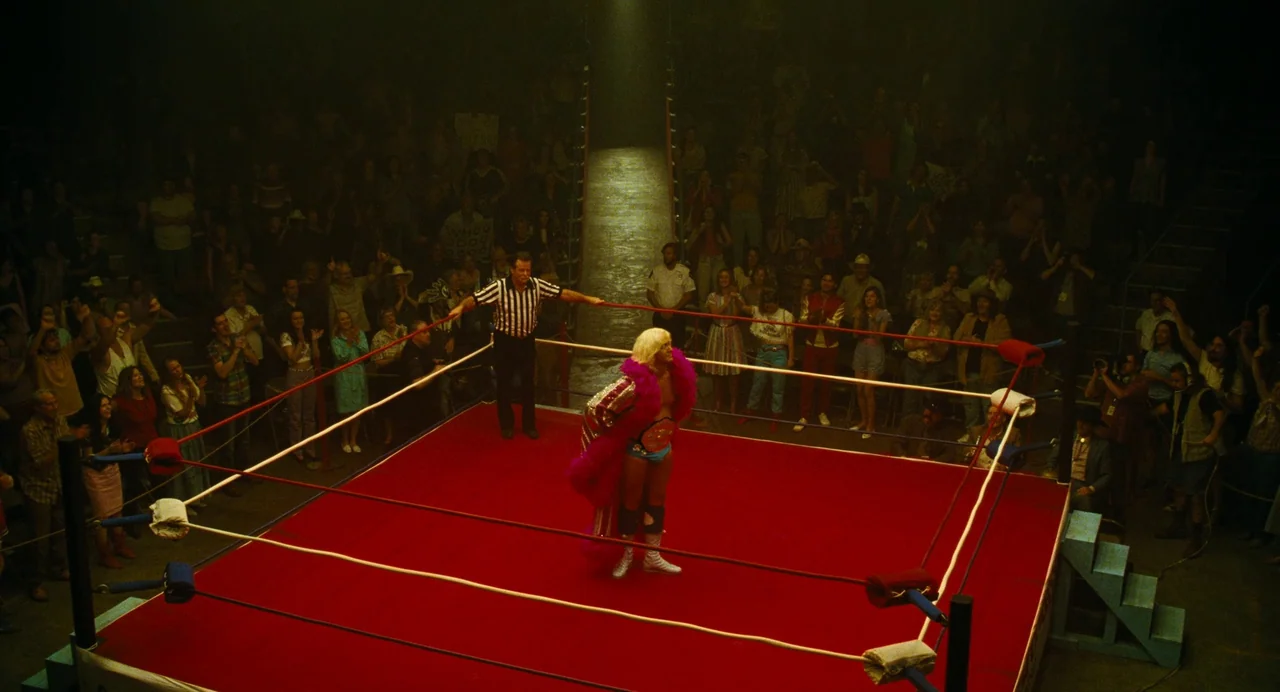 The Iron Claw (2023) movie still: wide — An indoor sports arena features a bright red wrestling ring surrounded by a seated audience. A blonde…